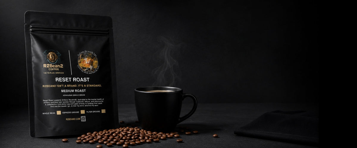 A bag of R2BEAN2 Coffee Reset Medium Roast stands beside a steaming ceramic mug of coffee, with roasted coffee beans scattered across a table. 